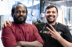 Zwei Mitarbeiter in der Küche; einer in weinroter Kochjacke, der andere in schwarzem Shirt und Schürze.