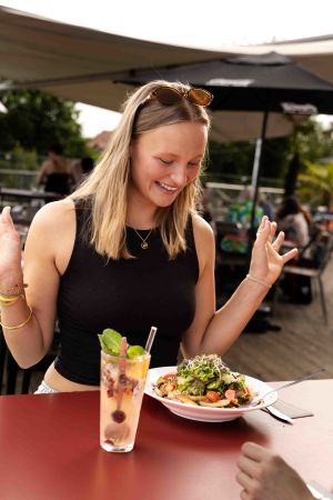 Eine junge Frau mit hochgeschobener Sonnenbrille sitzt an einem roten Tisch vor einem bunten Salat und einem Kaltgetränk.