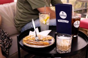 Eine Waffel mit Puderzucker auf einem Teller, zwei Kaltgetränke und eine blaue Speisekarte der „HERMANN Dachterrasse“ auf einem Tisch.