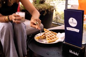 Eine Frau in lockerer Kleidung isst eine Waffel an einem Tisch, daneben steht eine „HERMANN Dachterrasse“-Speisekarte.