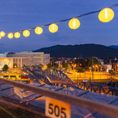 Blick von der Dachtereasse auf die Wiwilíbrücke und das Konzerthaus in Freiburg bei Dämmerung, eingerahmt von einer Lichterkette.