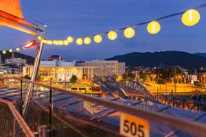 Blick von der Dachtereasse auf die Wiwilíbrücke und das Konzerthaus in Freiburg bei Dämmerung, eingerahmt von einer Lichterkette.