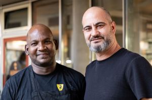 Zwei Männer in schwarzer Kleidung, einer davon trägt eine Schürze mit gelbem Logo.