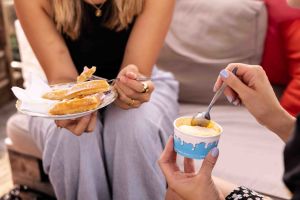 Zwei Frauen mit Desserts: Eine Person hält einen Teller mit Waffeln, die andere löffelt Eis aus einem blauen Pappbecher.
