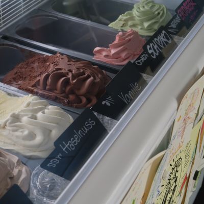 Blick in eine Eisvitrine mit verschiedenen Eissorten in Edelstahlbehältern. Schwarze Schildchen beschriften die Sorten wie „Haselnuss“, „Vanille“, „Schoko“ und „Erdbeer“.
