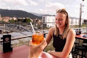Ein Aperol Spritz wird ins Bild gehalten und eine lächelnde junge Frau sitzt an einem Tisch auf der Dachterrasse.