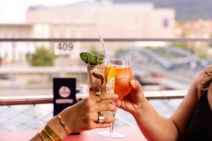 Zwei Hände stoßen mit Cocktails auf der Dachterrasse mit Stadtblick im Hintergrund an.