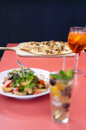 Ein roter Tisch, auf dem ein Flammkuchen auf einem Holzbrett, ein gemischter Salat und ein Kaltgetränk serviert sind.